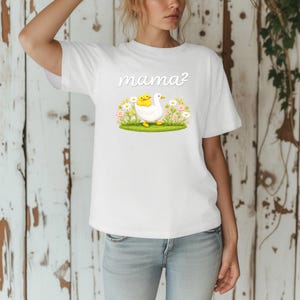 May include: White t-shirt with a graphic of a white goose carrying two yellow ducklings, set in a field of flowers and grass. The word "mama?" is written above the graphic in a cursive font. The shirt is worn by a person.