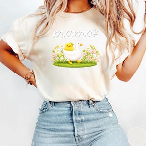 May include: A cream-coloured t-shirt with the text "mama&sup2;" above a graphic of a white duck carrying two yellow ducklings, set against flowers and grass. The shirt is made of a soft material.
