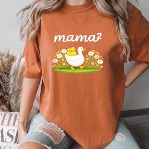 May include: Burnt orange t-shirt with the text "mama&sup2;" in white. The graphic shows a white duck with two yellow ducklings, set in a floral and grassy scene.