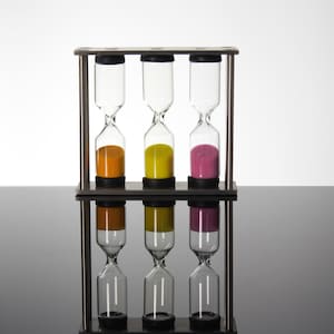 May include: A wooden-framed hourglass set with three glass hourglasses. Each hourglass contains a different colour of sand: orange, yellow, and pink. The hourglasses are arranged vertically in a dark wooden frame.