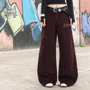 May include: Wide-leg burgundy corduroy pants with distressed details. Features a black belt with silver buckle and a small embroidered design on the pocket. The pants have a relaxed fit and a vintage aesthetic.