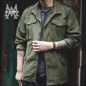 Op de afbeelding: Olijfgroene jas in militaire stijl met meerdere zakken en een ritssluiting. De jas heeft een kraag en lange mouwen. De mouwen zijn opgerold. De jas wordt gedragen over een donkergrijs t-shirt en een blauwe jeans. De jas heeft een logo aan de linkerbovenkant.