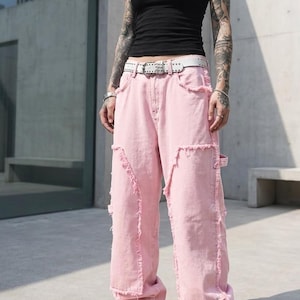Peut inclure: Un débardeur noir associé à un jean en denim rose clair et vieilli. Le jean présente des bords effilochés et une coupe large. Une ceinture blanche cloutée et des baskets blanches complètent la tenue.