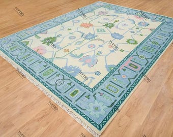 May include: Rectangular area rug with a cream-colored field and a light blue border. The rug features a floral pattern in shades of blue, pink, and green. The border has geometric and floral designs. The rug is placed on a wooden floor.
