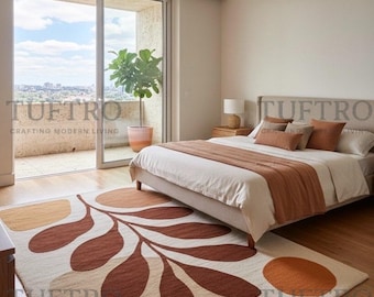 May include: A bedroom scene featuring a large area rug with a leaf and circle design in shades of brown, beige, and orange. The rug is placed in front of a bed with a neutral-colored headboard and bedding. The room has a balcony with a view.