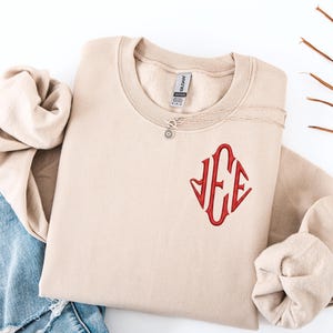 May include: A beige sweatshirt with a red embroidered monogram. The sweatshirt is folded with the sleeves tucked in. Light blue ripped jeans are next to the sweatshirt. A necklace with a small pendant is on the neckline.