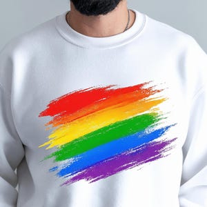 May include: White sweatshirt featuring a vibrant rainbow brushstroke design across the chest. The design includes red, orange, yellow, green, blue, and purple colors. The sweatshirt has a crew neck and long sleeves.