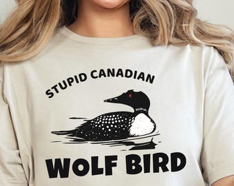 May include: Beige t-shirt with the text "STUPID CANADIAN WOLF BIRD" above and below a black and white illustration of a loon. The shirt is worn by a person sitting on a white couch.