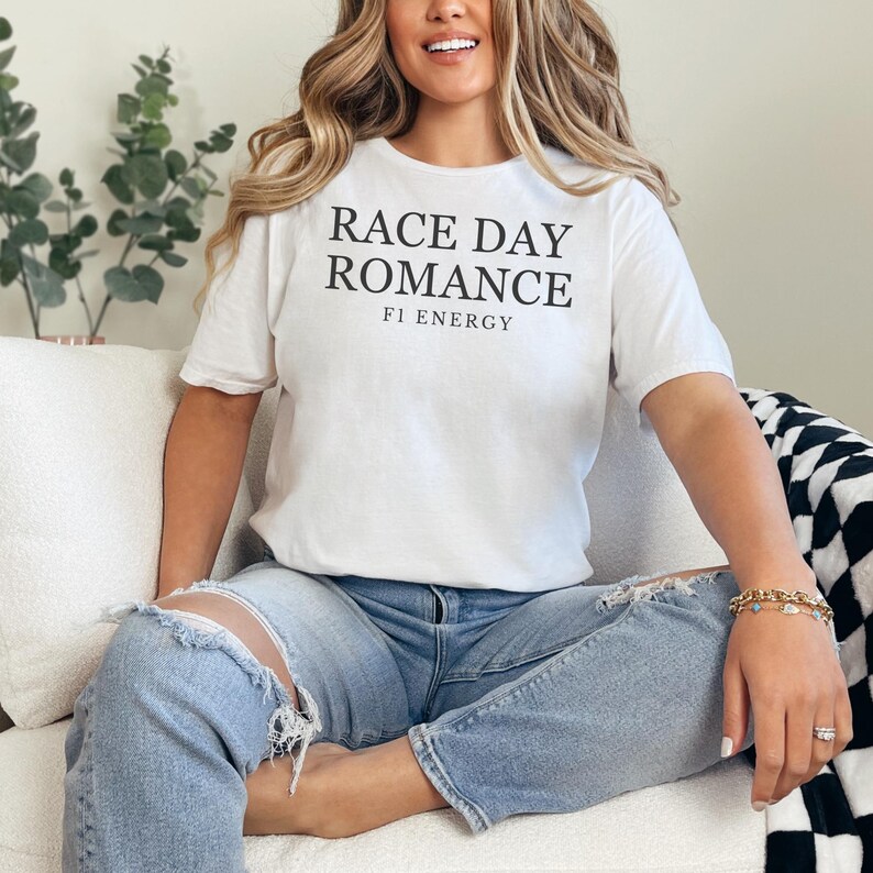 May include: White t-shirt with the text "RACE DAY ROMANCE" and "F1 ENERGY" in black. The shirt is paired with light blue ripped jeans. The person is sitting on a white couch.