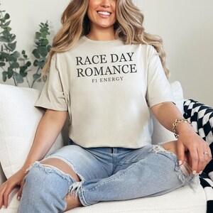 May include: A beige t-shirt with the text "RACE DAY ROMANCE" and "F1 ENERGY" in black. The shirt is paired with light blue ripped jeans. The model is sitting on a white couch.