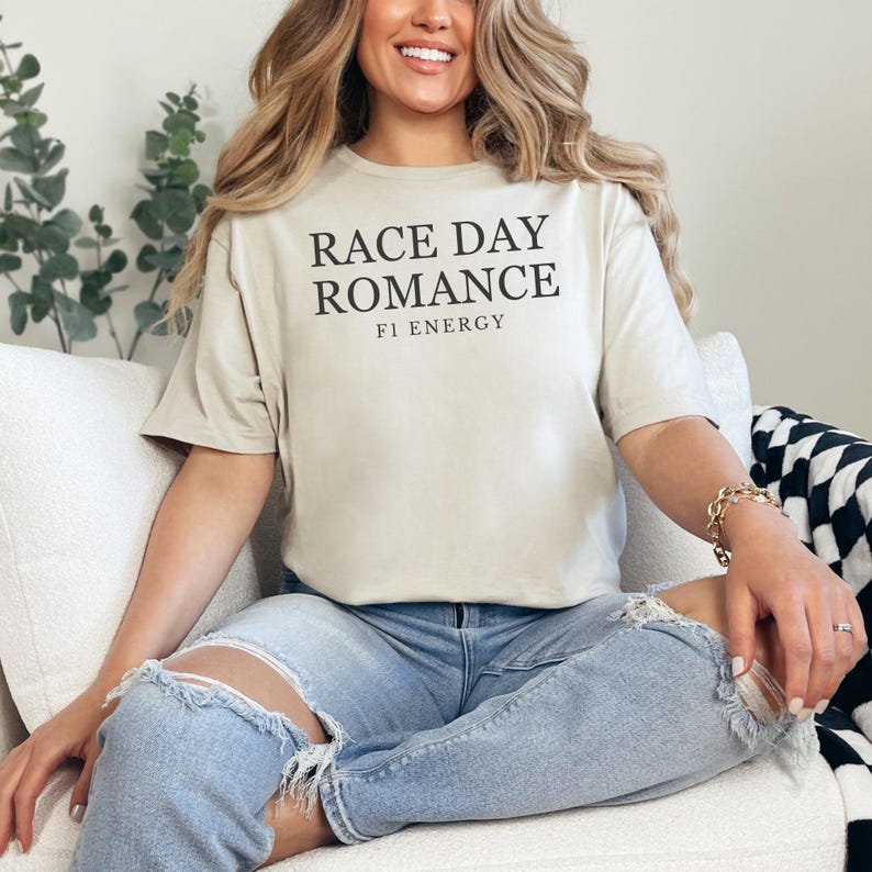 May include: A beige t-shirt with the text "RACE DAY ROMANCE" and "F1 ENERGY" in black. The shirt is paired with light blue ripped jeans. The model is sitting on a white couch.