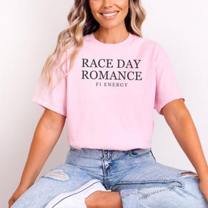 May include: A light pink t-shirt with the text "RACE DAY ROMANCE" and "F1 ENERGY" in black. The shirt is made of a soft material and has short sleeves.