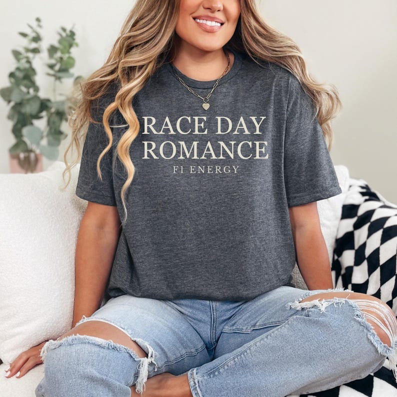 May include: Dark gray t-shirt with the text "RACE DAY ROMANCE" and "FI ENERGY" in white. The shirt is a crew neck style and appears to be made of a soft material. The model is wearing blue jeans.