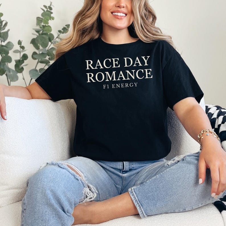 May include: Black t-shirt with the text "RACE DAY ROMANCE" and "F1 ENERGY" in white. The shirt is worn with light blue ripped jeans. The person is sitting on a white couch.