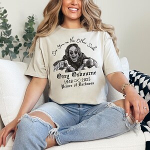 Może przedstawiać: Beżowa koszulka z czarno-białą grafiką Ozzy'ego Osbourne'a i napisem "See You on the Other Side". Poniżej znajduje się "Ozzy Osbourne 1948 &infin; 2025 Prince of Darkness" w gotyckiej czcionce. Koszulka jest noszona.