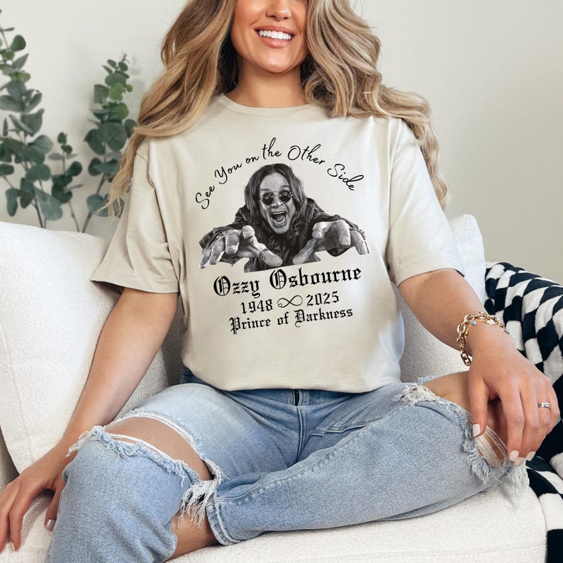 Może przedstawiać: Beżowa koszulka z czarno-białą grafiką Ozzy'ego Osbourne'a i napisem "See You on the Other Side". Poniżej znajduje się "Ozzy Osbourne 1948 &infin; 2025 Prince of Darkness" w gotyckiej czcionce. Koszulka jest noszona.