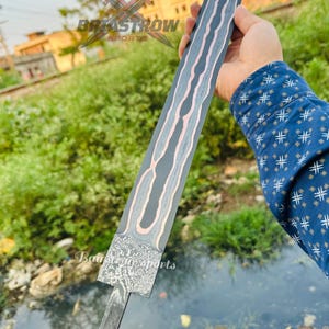 May include: A long, straight sword blade featuring a dark, patterned Damascus steel finish with a prominent, wavy copper-colored inlay running down the center. The base of the blade has a textured, mottled metal surface and a notched tang extension.