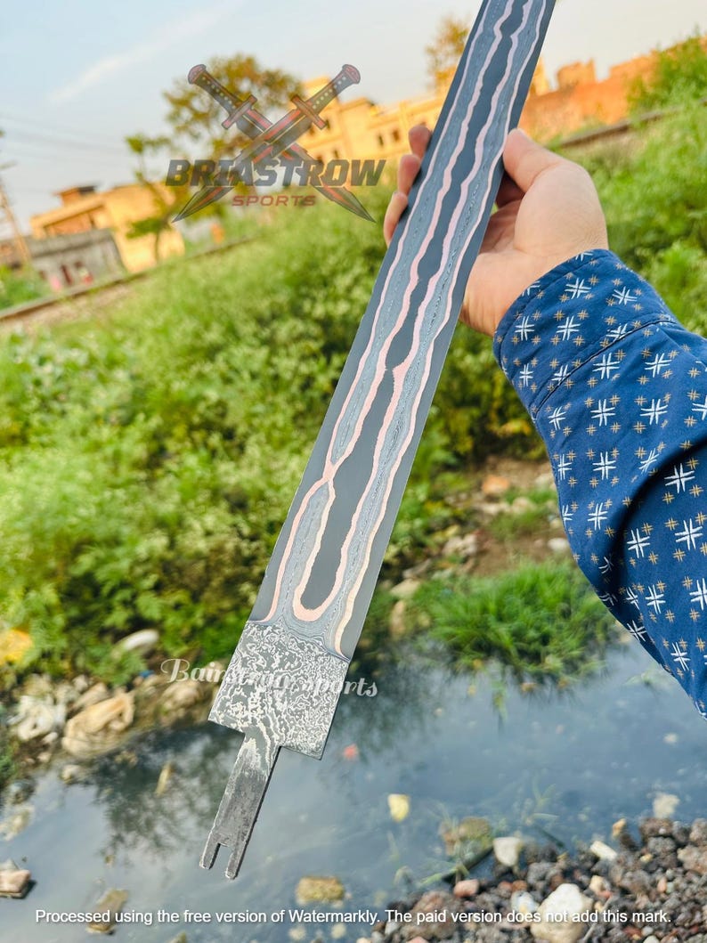 May include: A long, straight sword blade featuring a dark, patterned Damascus steel finish with a prominent, wavy copper-colored inlay running down the center. The base of the blade has a textured, mottled metal surface and a notched tang extension.