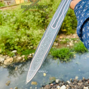 May include: A double-edged sword blade featuring a central panel with a complex, swirling Damascus steel pattern, bordered by smooth, light-colored metal edges. The blade displays the text Briastrow Sports and Bairstrow sports.