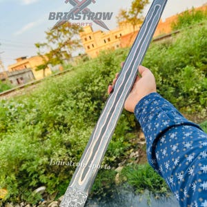 May include: A long, double-edged sword blade featuring a dark, textured finish with prominent, wavy copper-colored inlay lines running along the length of the blade. The base of the blade displays a rough, hammered metal pattern and a tang extension.
