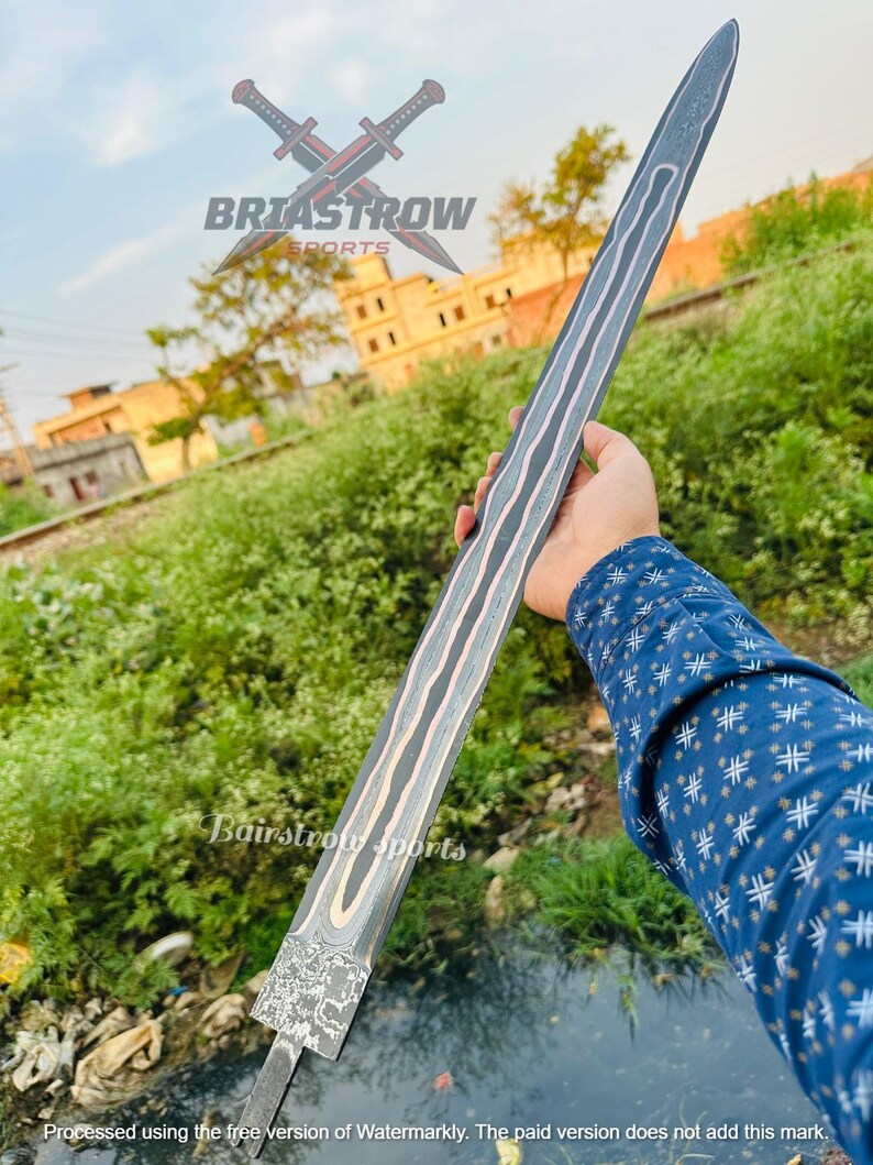 May include: A long, double-edged sword blade featuring a dark, textured finish with prominent, wavy copper-colored inlay lines running along the length of the blade. The base of the blade displays a rough, hammered metal pattern and a tang extension.