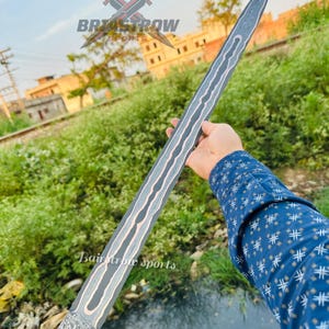 May include: A long, double-edged sword blade featuring a dark, textured metal finish with a prominent, wavy copper-colored inlay running down the center. The base of the blade displays a complex, mottled pattern, and the tip is pointed.