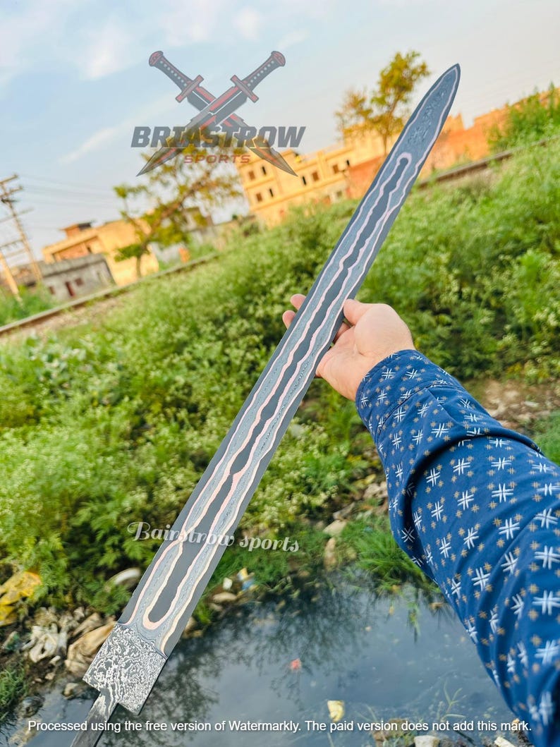May include: A long, double-edged sword blade featuring a dark, textured metal finish with a prominent, wavy copper-colored inlay running down the center. The base of the blade displays a complex, mottled pattern, and the tip is pointed.