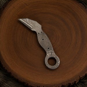 May include: A Damascus steel knife blade blank featuring a distinct wavy pattern on the metal surface. The blade has a sharp, angled tip, a curved spine, and a handle section with three small circular holes and a large finger ring at the end.