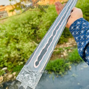 May include: A double-edged sword blade featuring a dark gray finish with a prominent, wavy, light-colored Damascus steel pattern running down the center. The base of the blade displays a textured, mottled metal surface with a distinct rectangular transition.
