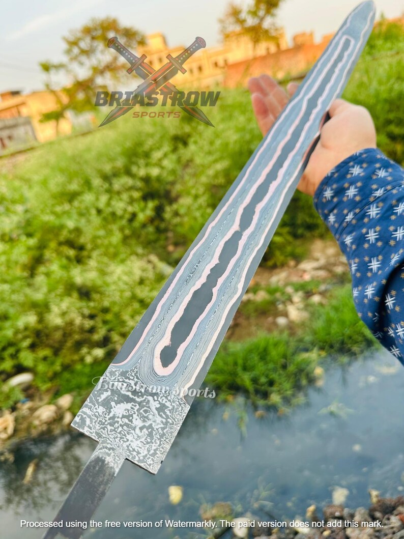 May include: A double-edged sword blade featuring a dark gray finish with a prominent, wavy, light-colored Damascus steel pattern running down the center. The base of the blade displays a textured, mottled metal surface with a distinct rectangular transition.