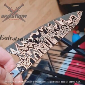 May include: A metal knife blade featuring a distinct wavy, multi-toned pattern of black, grey, and cream colors. The blade has a pointed tip and a curved edge. The text BRIASTROW SPORTS and Bairstrow sports are visible on the blade surface.