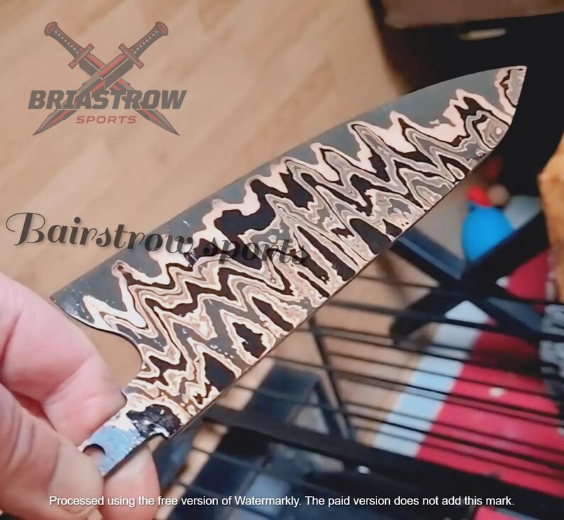 May include: A metal knife blade featuring a distinct wavy, multi-toned pattern of black, grey, and cream colors. The blade has a pointed tip and a curved edge. The text BRIASTROW SPORTS and Bairstrow sports are visible on the blade surface.