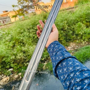 May include: A long, straight sword blade featuring a dark, layered Damascus steel pattern with a prominent, wavy central fuller. The metal displays a distinct contrast between dark grey and lighter silver tones, with a visible tang at the top end.