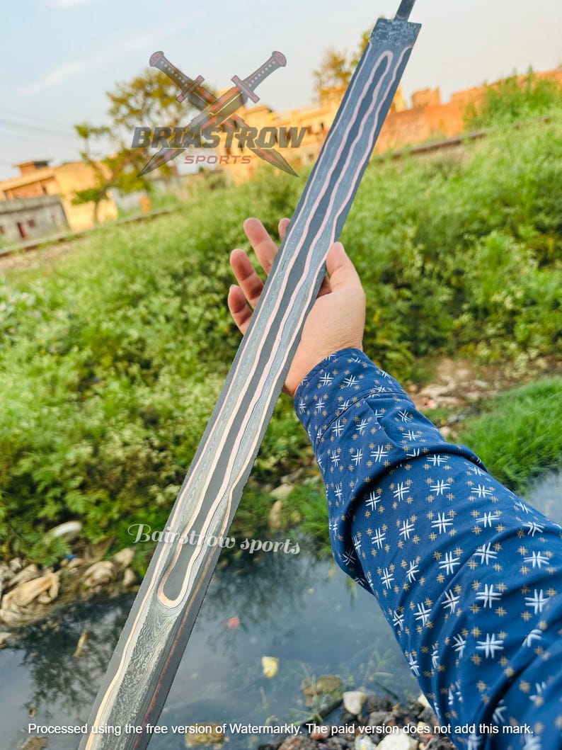May include: A long, straight sword blade featuring a dark, layered Damascus steel pattern with a prominent, wavy central fuller. The metal displays a distinct contrast between dark grey and lighter silver tones, with a visible tang at the top end.