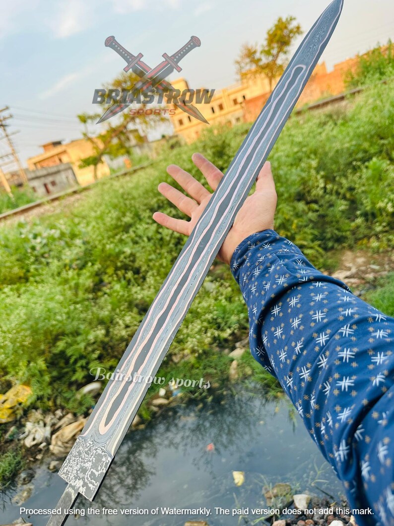 May include: A long, straight sword blade featuring a distinct Damascus steel pattern with wavy, light-colored metallic lines running along the length of the blade. The base of the blade displays an intricate, etched design of a roaring lion's head.