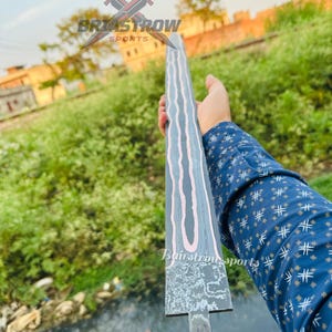 May include: A long, straight sword blade featuring a dark, textured finish with prominent, wavy, light-colored lines running vertically along its length. The blade is held horizontally, displaying a distinct pattern and a flat, unsharpened tang base.