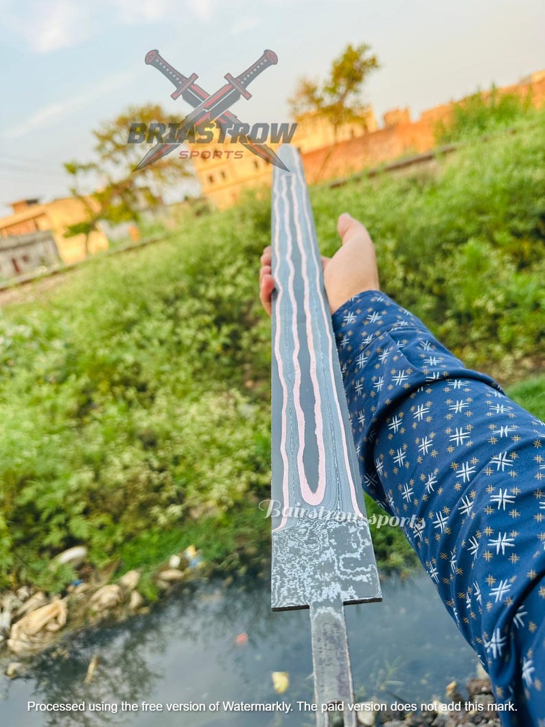 May include: A long, straight sword blade featuring a dark, textured finish with prominent, wavy, light-colored lines running vertically along its length. The blade is held horizontally, displaying a distinct pattern and a flat, unsharpened tang base.