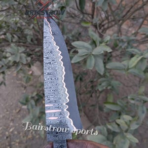 Può includere: Una lama di coltello in acciaio Damasco con un design a motivi e un bordo nero. La lama ha un bordo seghettato e un accento color rame. Il testo "Bairstrow Sports" è visibile sulla lama.