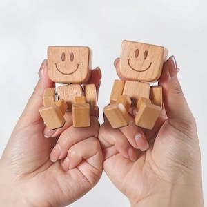 以下が含まれることがあります： 笑顔の顔が付いた2つの木製関節人形。各人形は正方形の頭、長方形の胴体、関節のある腕と脚を持っています。人形は明るい色の木でできており、白い背景に対して手で持たれています。