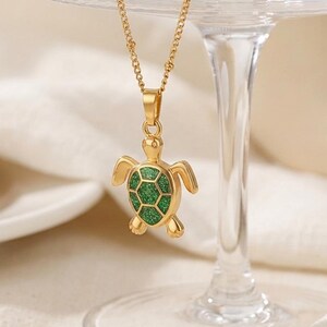 May include: A gold-coloured necklace featuring a turtle pendant. The pendant has a green, glittery shell and hangs from a fine chain. The necklace is shown against a soft, neutral backdrop, emphasising the jewellery's design.