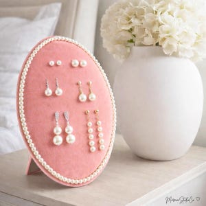 May include: A pink oval earring display with a pearl border holds various pairs of earrings. The earrings include small studs, dangling pearl earrings, and earrings with crystal accents. A white vase with white flowers sits to the right.