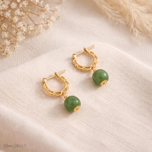 May include: A pair of gold-colored hoop earrings with a decorative openwork design. Each earring features a round, green gemstone pendant. The earrings are displayed on a white, textured surface.