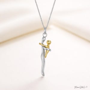 Op de afbeelding: Een zilveren en gouden hangerketting met twee figuren die elkaar omarmen. De grotere figuur is zilver, de kleinere goud, met armen om de zilveren figuur. De ketting hangt aan een fijne zilveren ketting.
