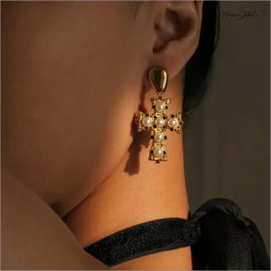 Può includere: Orecchini a croce dorati con perle e dettagli blu scuro. Il design a croce presenta una parte superiore a forma di goccia e un motivo ornato. Gli orecchini sono mostrati sull'orecchio e sul collo di una persona.