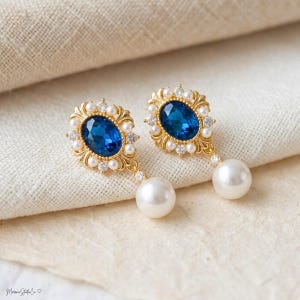 May include: A pair of gold earrings featuring a central oval sapphire-blue stone, surrounded by small pearls and crystals. Each earring has a large, dangling white pearl. The earrings are displayed on a cream-coloured fabric.