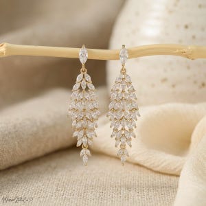 May include: A pair of gold-toned dangle earrings. Each earring features a cascade of clear, marquise-cut crystals, creating a leaf-like design. The earrings are suspended from a single, larger crystal at the top. The earrings are displayed on a light-colored wooden stick.