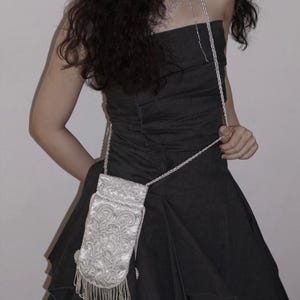 May include: A small, rectangular, silver-toned crossbody bag featuring intricate, raised floral embroidery and a decorative fringe along the bottom edge. The bag is suspended by a matching silver-toned chain strap, creating a structured accessory.
