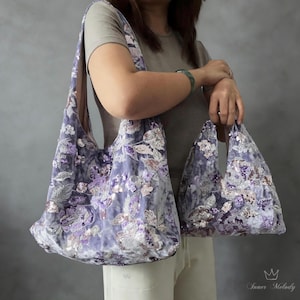 Pode incluir: Uma bolsa de ombro grande estilo hobo e uma bolsa de mão menor a combinar, ambas feitas de tecido de veludo lavanda. As bolsas apresentam um intrincado bordado floral com lantejoulas e contas prateadas e roxas. O texto Inner Melody é visível.