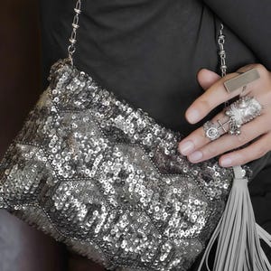May include: A silver sequinned handbag featuring a geometric quilted pattern, a metallic chain strap, and a long grey tassel attached to the side. The bag is covered in small, reflective silver sequins that create a textured, shimmering surface.