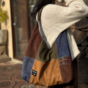May include: A patchwork shoulder bag in shades of brown, blue, and green. The bag features a variety of textures and patterns, including suede and plaid. A small label is visible on the front. The bag is carried over the shoulder.
